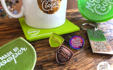 Three Made by Cooper pin badges surrounded by coffee cups, drinks mats and custom patches.