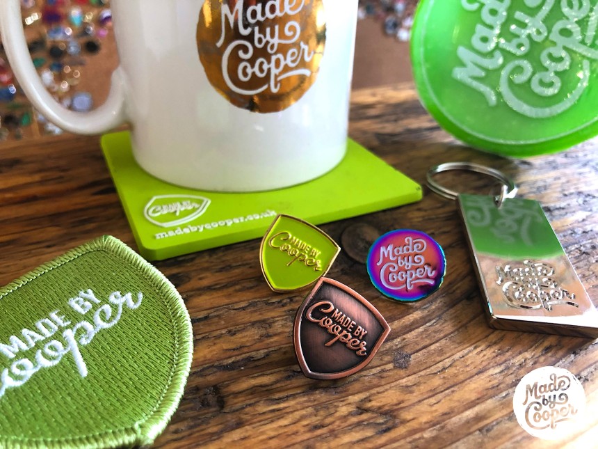Three Made by Cooper pin badges surrounded by coffee cups, drinks mats and custom patches.