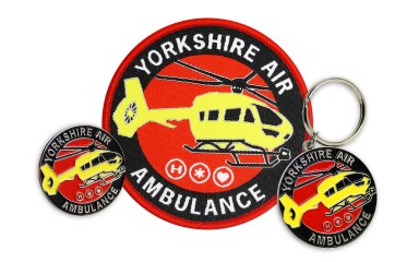 A collection of Yorkshire Air Ambulance merch. A small soft enamel pin badge, a chunky soft enamel keyring, and a large woven patch, all sporting the new YAA red, black, and yellow logo and branding.
