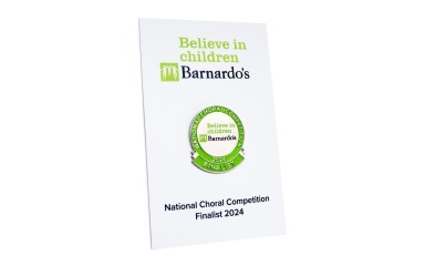 A Barnardo's special edition pin badges to commemorate the National Choral Competition of 2024. The green and silver pin badge is fastened to a white backing card with green and black text.