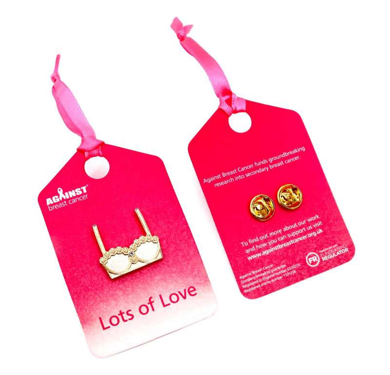 A golden bra enamel pin badge on a pink backing card with a pink ribbon attached to raise money for charity.