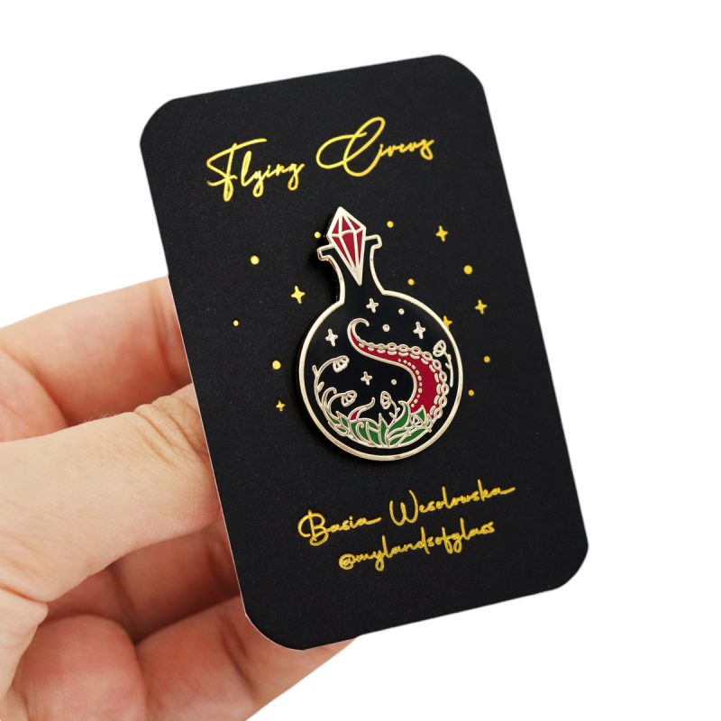 A weird hand holding an enamel pin that's connected to a backing card. The badge looks like a potion bottle with a dragon inside and the bottle neck is sealed by the reddest ruby ever.