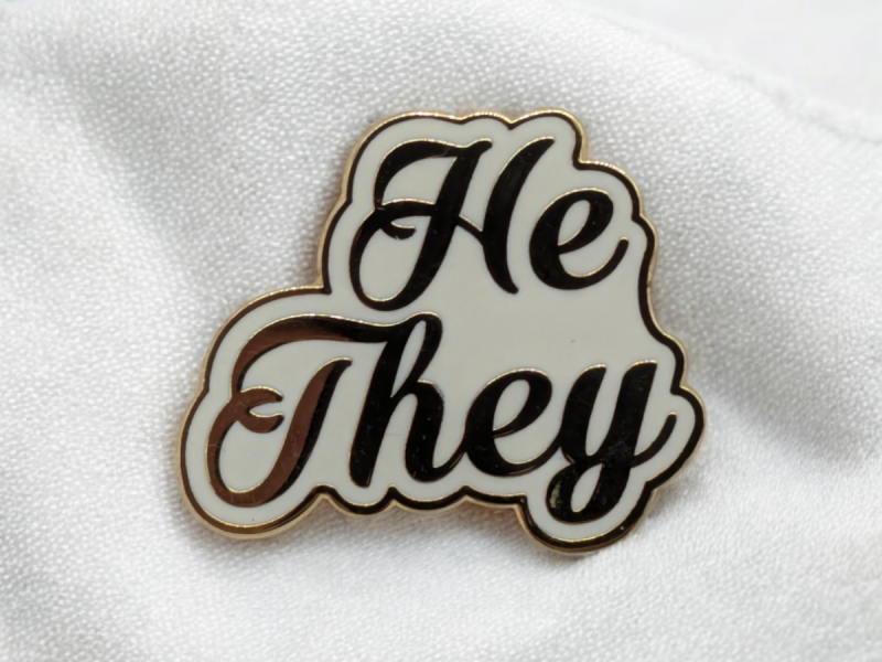 A He/They pronoun badge in a sweeping and swirling font pinned to some white fabric.