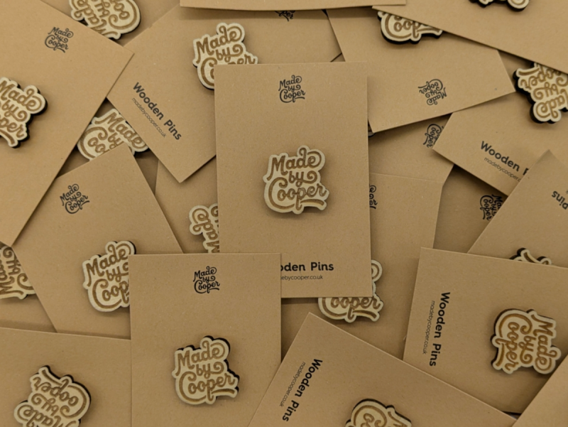 A pile of wooden pin badges of Made by Cooper logos and brown recyclable backing cards.