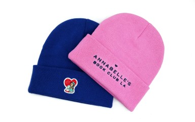 A pink and blue custom beanie. The blue hat features a love heart logo while the pink hat has 