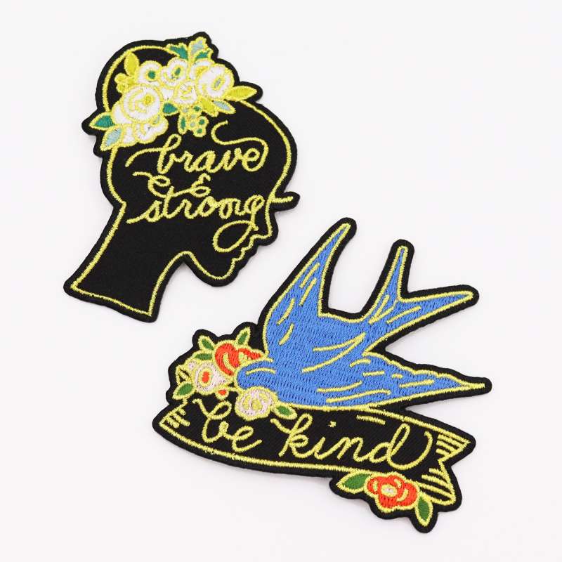 Two beautifully designed patches. One the silhouette of a woman's head, she has flowers in the her hair and the words 'Brave & Strong' are embroidered in stunning yellow thread. The second patch is a pale blue swallow with a banner that reads 'Be Kind'.