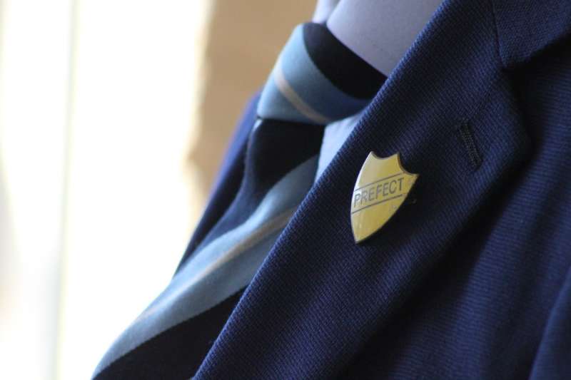 A close up of a blue school blazer with a yellow Prefect shield pinned to the lapel.