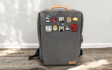 A grey backpack covered in brightly coloured pin badges sits on a wooden floor against a white wall backdrop. The sun creates a warm feeling around the bag.