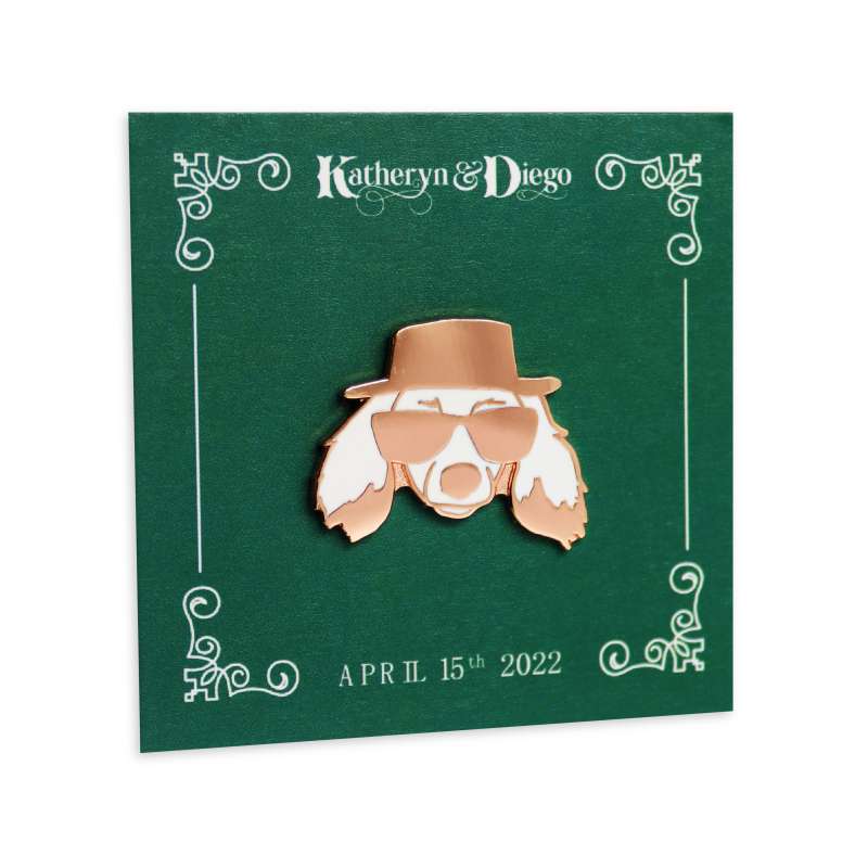 A funky dog pin badge. The dog has long floppy ears and is wearing sunglasses and a pork pie hat. The badge is attached to a green backing card with the names of the bride and groom and the date of the wedding.