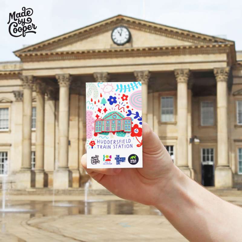 A stylised enamel pin of Huddersfield train station on a brightly coloured backing card being held up outside Huddersfield train station.