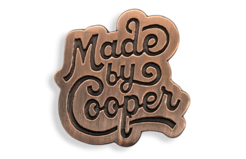 A bronze pin pin badge featuring the Made by Cooper logo.